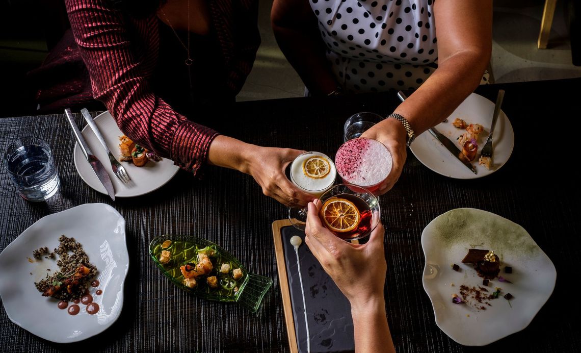 3 hands toast cocktails above a selection of dishes